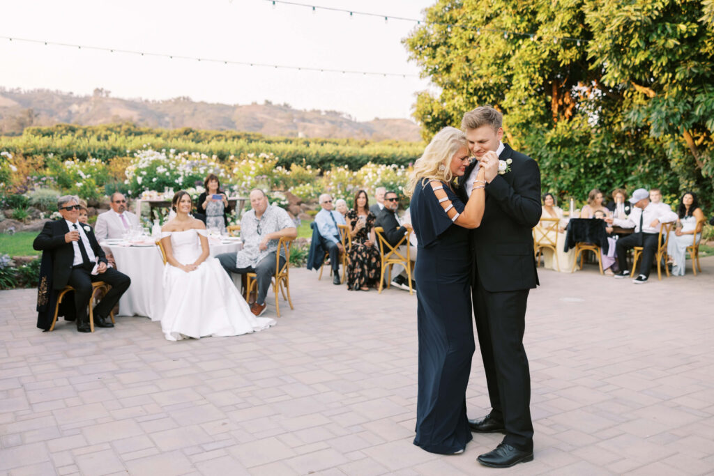 A groom in a black suit shares a joyful dance with a woman in a navy dress at an outdoor wedding reception, while guests seated at tables watch and smile in the background. Copyright - Lisa Leanne Photography, California
