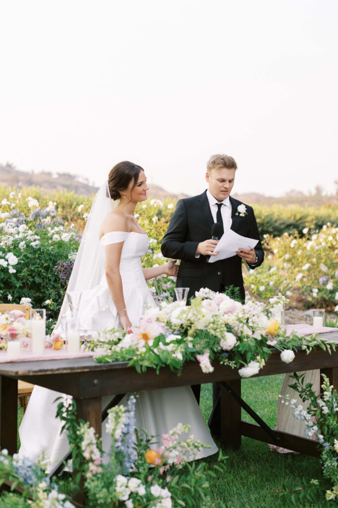 A bride in an off-the-shoulder white gown and a groom in a black suit stand behind a flower-adorned table outdoors. The groom holds a microphone and paper, appearing to give a speech. The setting is surrounded by greenery and flowers. Copyright - Lisa Leanne Photography, California