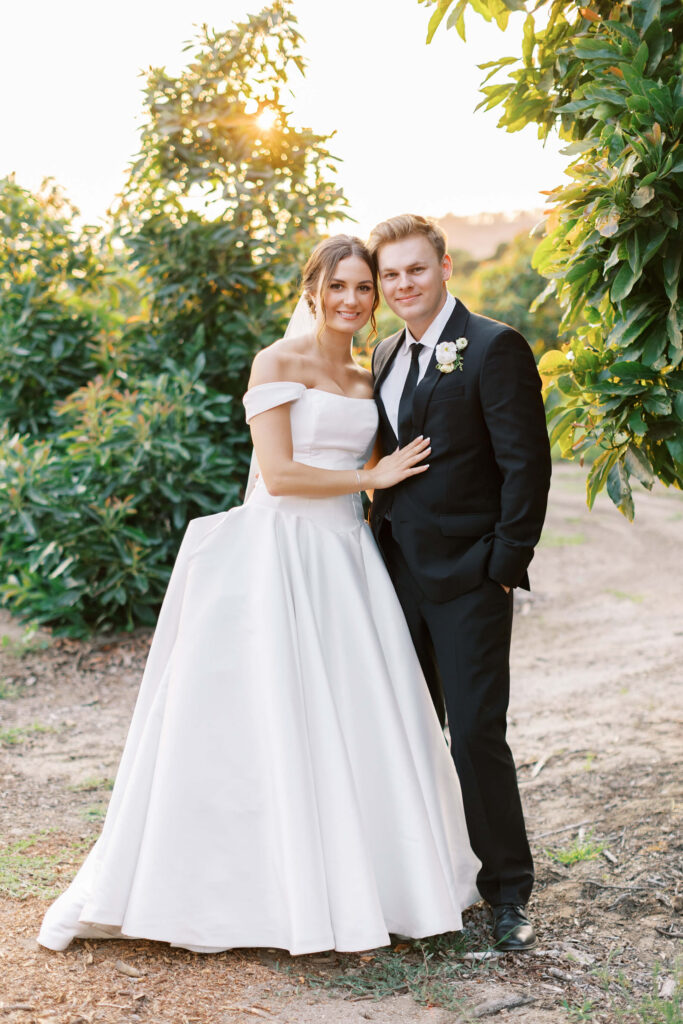 A bride in an off-the-shoulder white gown and a groom in a black suit with a boutonniere stand close together outdoors, surrounded by greenery and sunlight. Copyright - Lisa Leanne Photography, California