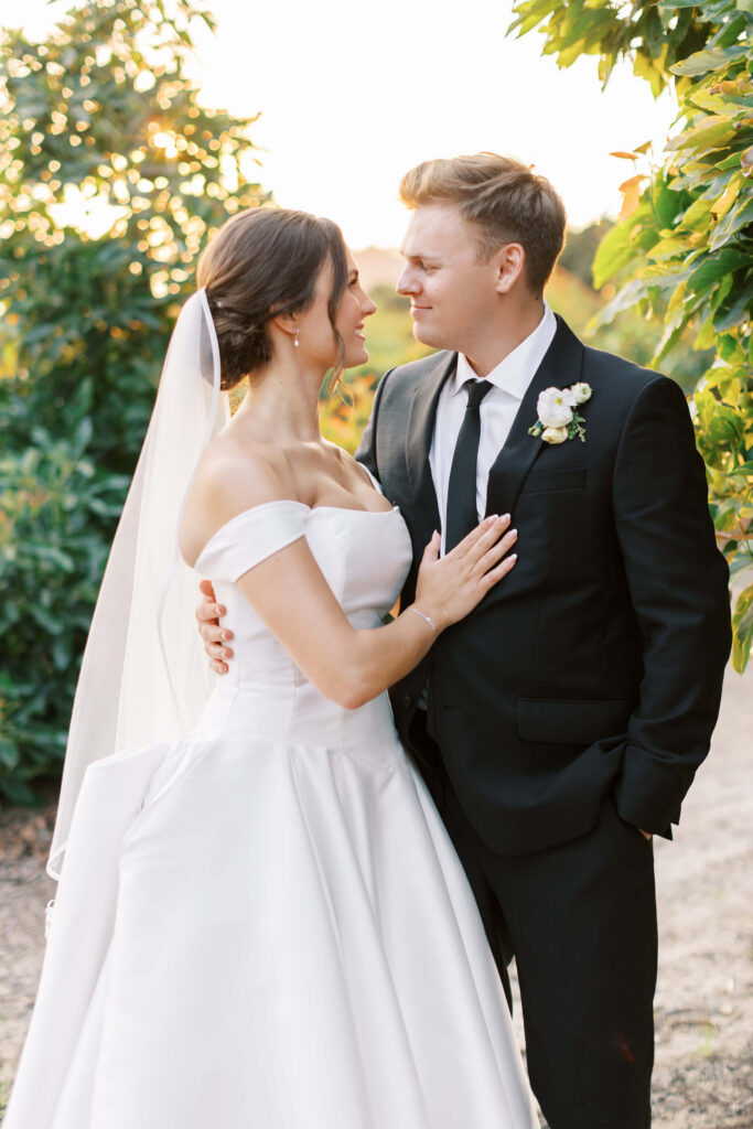 A bride in a white off-the-shoulder gown and veil stands closely with a groom in a black suit and tie. They gaze at each other lovingly outdoors, surrounded by greenery and soft sunlight. Copyright - Lisa Leanne Photography, California