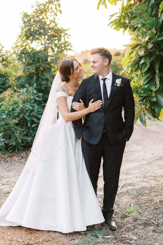 A bride in a white gown and veil embraces a groom in a black suit and tie outdoors, both smiling warmly at each other amid green trees and sunlight. Copyright - Lisa Leanne Photography, California