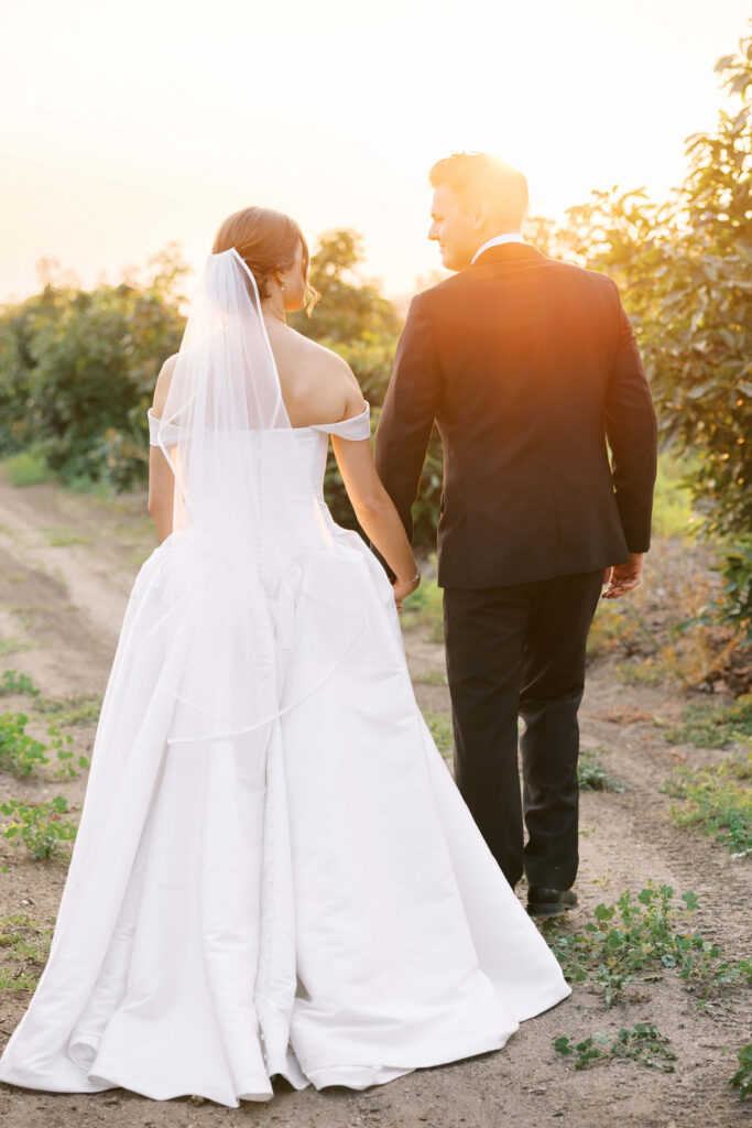 A bride in a white gown and veil holds hands with a groom in a black suit as they walk down a sunlit outdoor path surrounded by greenery, with the warm glow of sunset in the background. Copyright - Lisa Leanne Photography, California