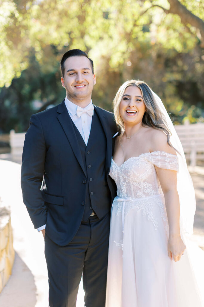 A smiling bride in an off-shoulder white wedding dress and veil stands next to a groom in a dark suit and white bow tie, outdoors in sunlight with trees and benches in the background. Copyright - Lisa Leanne Photography, California