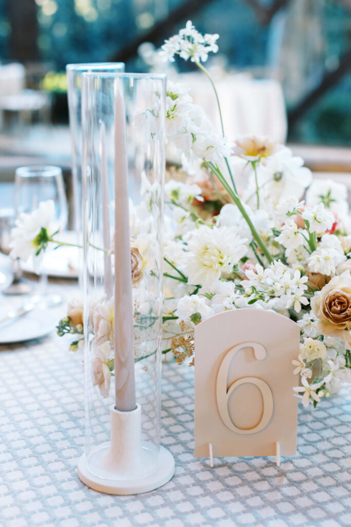 A table centerpiece with a tall white candle in a glass holder, a floral arrangement of white and pale yellow flowers, and a table number card displaying the number 6 on a patterned tablecloth. Copyright - Lisa Leanne Photography, California