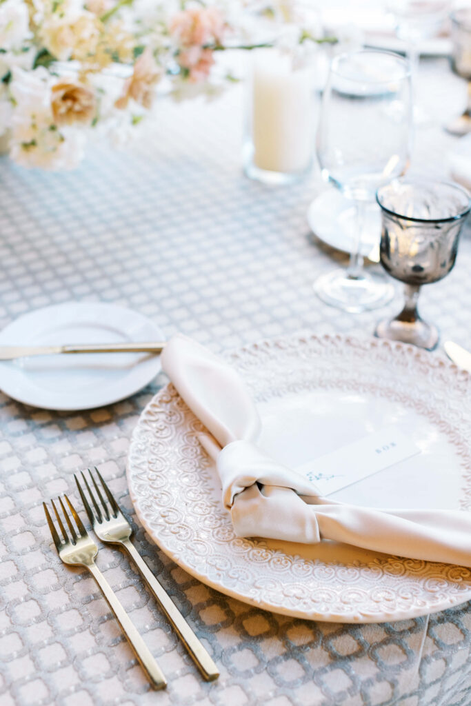 Oak Room Reception at Calamigos Ranch Elegant table setting with a decorative plate, neatly folded napkin, name card, gold utensils, wine glass, water glass, and a small plate with a knife. Soft floral arrangement and candle in the background. Copyright - Lisa Leanne Photography, California