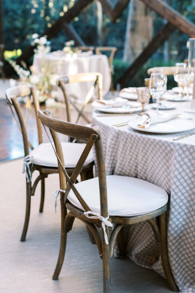 Calamigos Ranch Wedding Wooden chairs with white cushions are arranged around a round table covered with a patterned white tablecloth, set with glassware, plates, and napkins in an elegant, sunlit setting. Copyright - Lisa Leanne Photography, California
