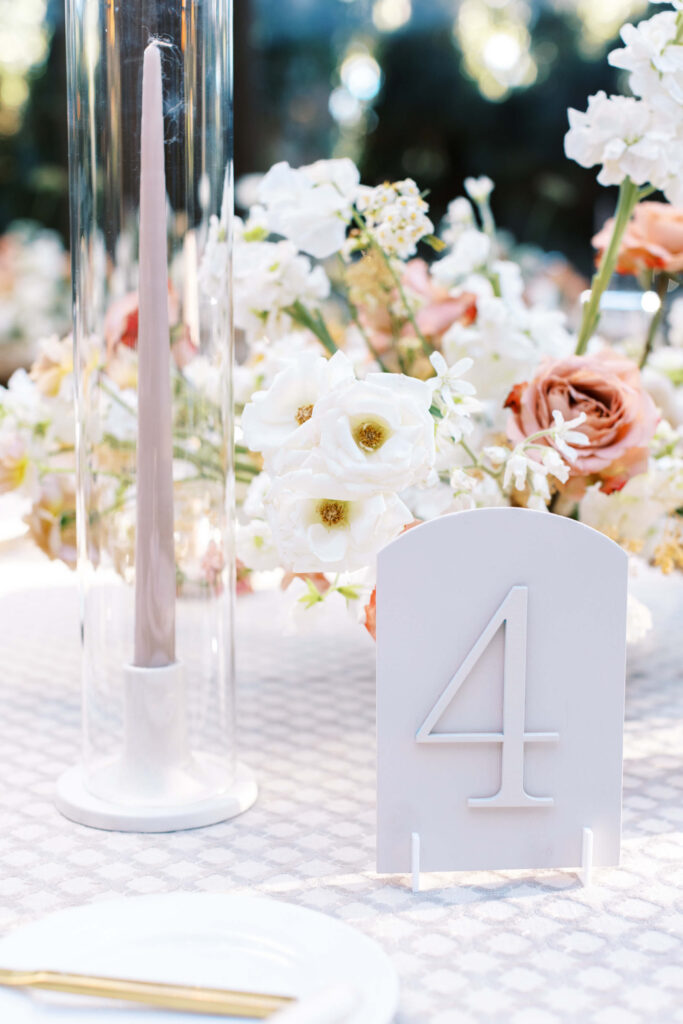 Oak Room Reception white table number card with the number 4 stands on a patterned tablecloth next to a tall, pale taper candle in a glass holder and a floral arrangement of white and blush flowers. Copyright - Lisa Leanne Photography, California