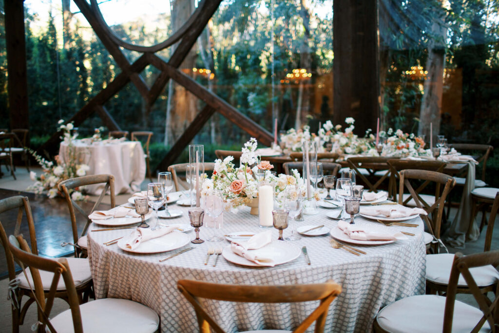 Oak Room Wedding Elegant reception setup with round tables covered in white linens, surrounded by wooden chairs. Floral centerpieces, candles, and glassware decorate the tables. Large windows reveal lush greenery outside. Copyright - Lisa Leanne Photography, California