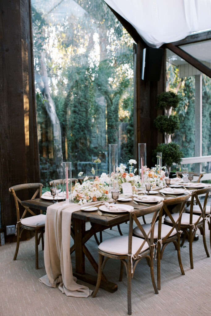 Calamigos Ranch Oak Room dining table set for six with white plates, glassware, beige napkins, candle holders, and pastel floral centerpieces, beneath a clear tent with views of trees and greenery outside. Copyright - Lisa Leanne Photography, California
