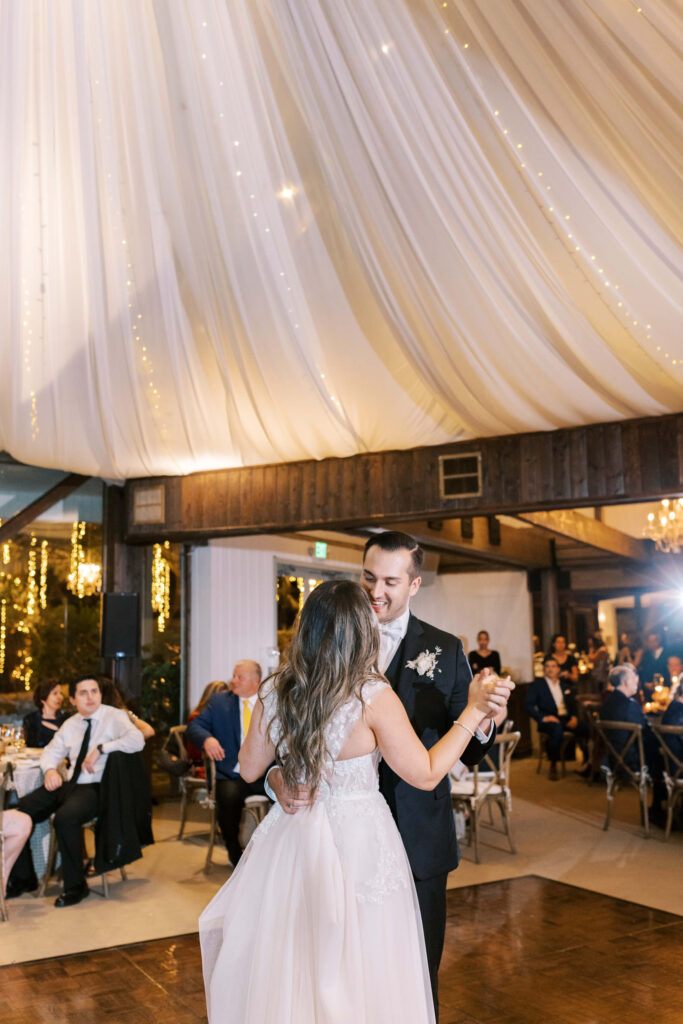 Oak Room Reception Bride and Groom first dance under a white draped ceiling with string lights, while guests seated at tables watch and celebrate in the warmly lit venue. Copyright - Lisa Leanne Photography, California