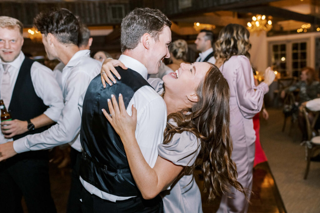 A joyful couple dances closely at a Calamigos Ranch Oak Room Reception, smiling and laughing together while people mingle and dance in the warmly lit background. Copyright - Lisa Leanne Photography, California
