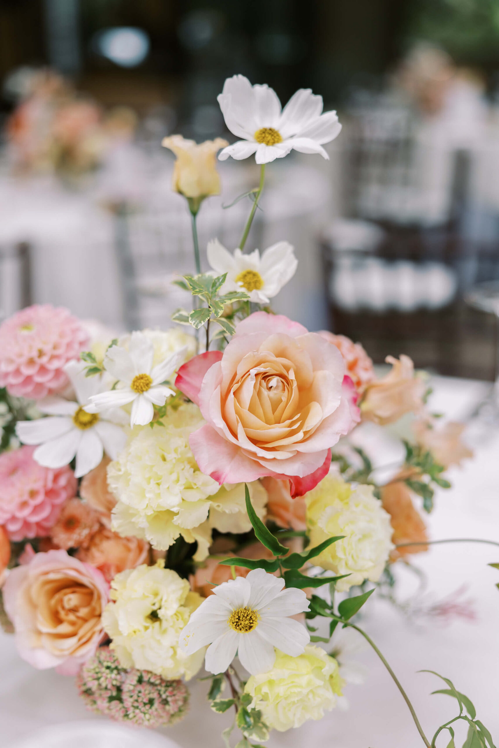 A floral arrangement featuring pale pink and peach roses, white daisies, yellow carnations, and assorted greenery, set on a white tablecloth with blurred chairs and tables in the background. Copyright - Lisa Leanne Photography, California