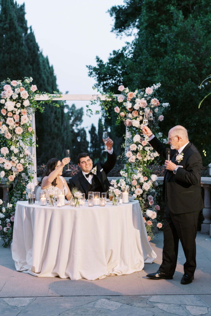 Villa Del Sol D'Oro Reception Toast  - A bride and groom sit at a round table decorated with flowers and candles, raising glasses in a toast as an older man in a tuxedo stands beside them, also toasting, under a floral arch at an outdoor wedding reception. Copyright - Lisa Leanne Photography, California