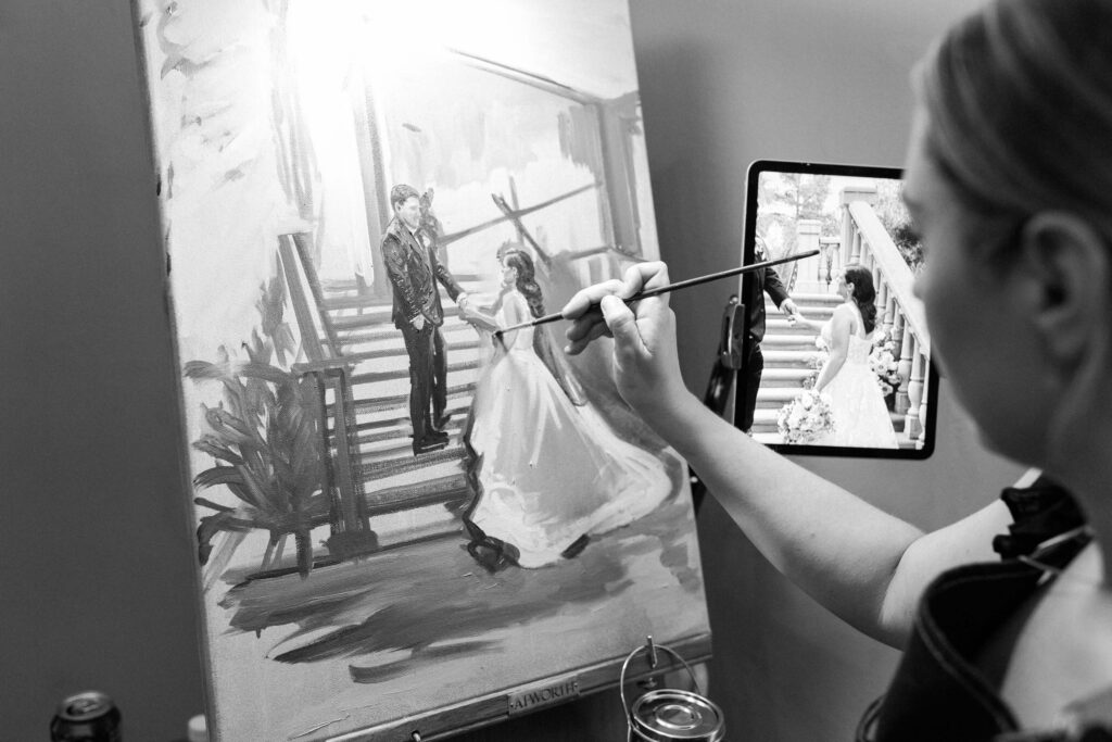 Villa Del Sol D'Oro Wedding Live Painter - A woman paints a wedding scene on a canvas, using a reference photo of a bride and groom on stairs displayed on a tablet next to her. The scene is depicted in black and white. Copyright - Lisa Leanne Photography, California