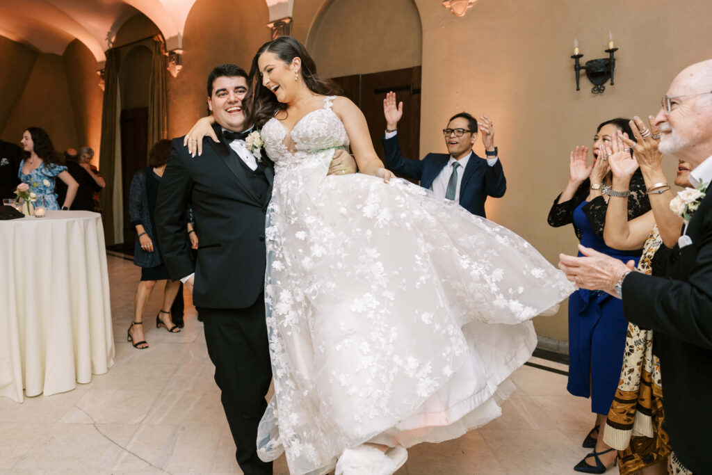 Villa Del Sol D'Oro Reception  - A smiling groom in a black tuxedo carries his laughing bride, who wears a white floral wedding dress, as wedding guests cheer and celebrate around them indoors. Copyright - Lisa Leanne Photography, California