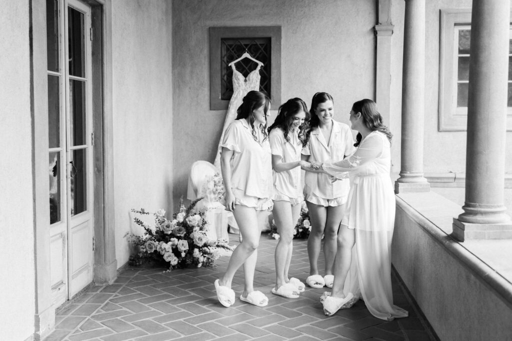 Villa Del Sol D'Oro Wedding - Four women in matching pajamas and slippers smile and hold hands on a veranda. A wedding dress hangs in the background above a floral arrangement. The scene is joyful and relaxed. Copyright - Lisa Leanne Photography, California