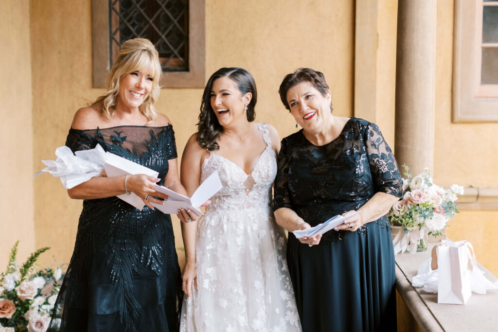 Villa Del Sol D'Oro Wedding - Three women dressed formally, including a bride in a white gown, stand smiling and laughing together while holding white gift boxes. They are indoors, surrounded by flowers and soft natural light. Copyright - Lisa Leanne Photography, California