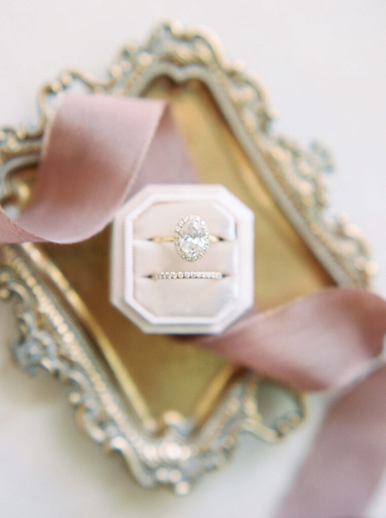 Villa Del Sol D'Oro Wedding Bridal Details - An elegant engagement ring and wedding band set with diamonds in a white velvet ring box, placed on a decorative gold tray with a soft blush pink ribbon. Copyright - Lisa Leanne Photography, California