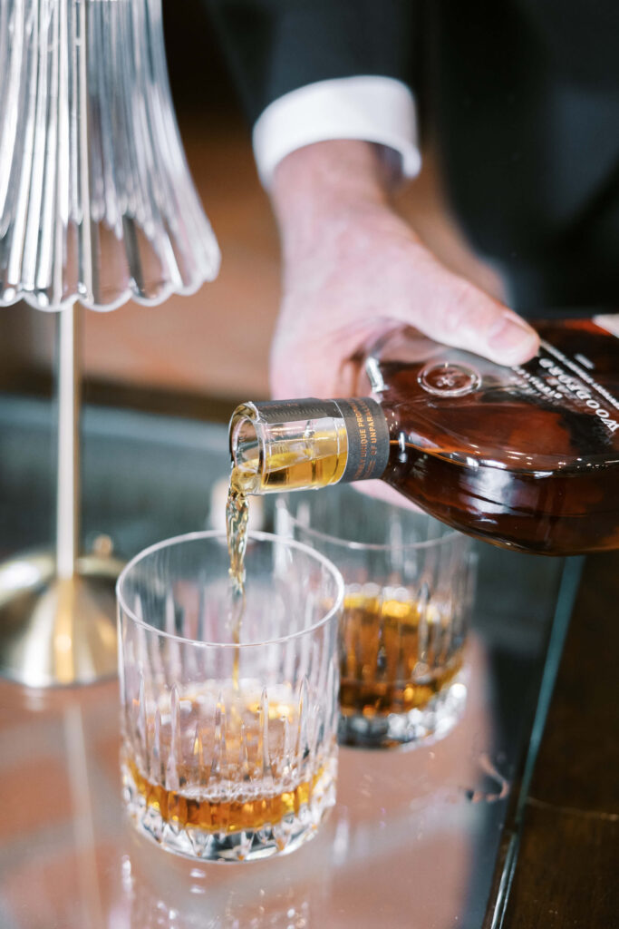 Villa Del Sol D'Oro Wedding - A person pours whiskey from a brown bottle into a crystal glass on a reflective table, with another filled glass and a decorative lamp nearby. Copyright - Lisa Leanne Photography, California