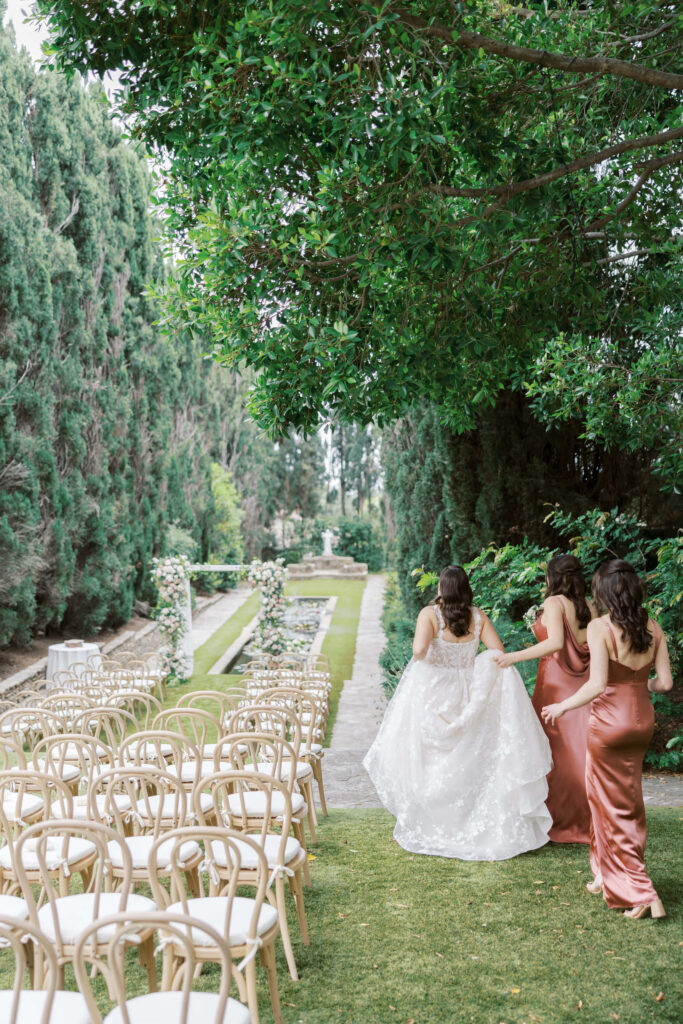 Villa Del Sol D'Oro Wedding -Three women walk down a grassy aisle lined with empty chairs and trees. One wears a white wedding dress, and the other two wear matching pink dresses, holding the bride’s train as they approach a floral wedding arch. Copyright - Lisa Leanne Photography, California