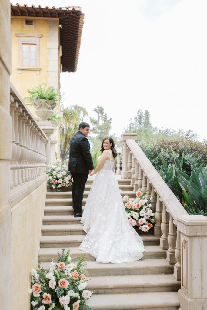 Bride and Groom Portrait at Villa Del Sol D'Oro - A bride and groom stand on stone steps, holding hands and looking back at the camera. The bride wears a white gown, the groom a black suit. The steps are decorated with pink and white flowers, and a villa is in the background. Copyright - Lisa Leanne Photography, California