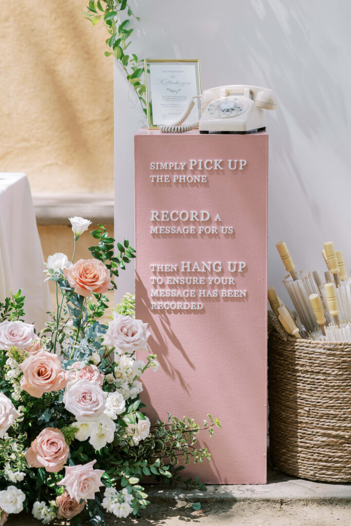 Villa Del Sol D'Oro Wedding Ceremony - A pink pedestal with instructions to pick up a vintage phone, record a message, and hang up. Surrounded by a floral arrangement, a framed sign, and a basket of white umbrellas. Copyright - Lisa Leanne Photography, California