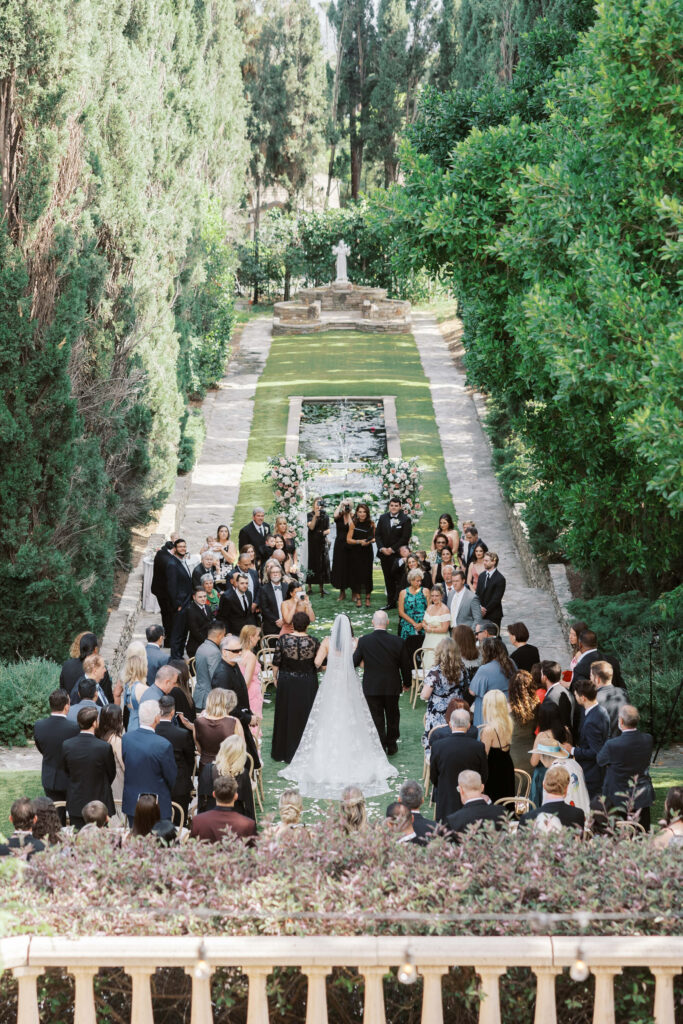 Villa Del Sol D'Oro Wedding Ceremony - A bride in a white dress walks down an outdoor aisle lined with guests toward a floral wedding arch, surrounded by lush greenery and tall trees in a garden setting. Copyright - Lisa Leanne Photography, California