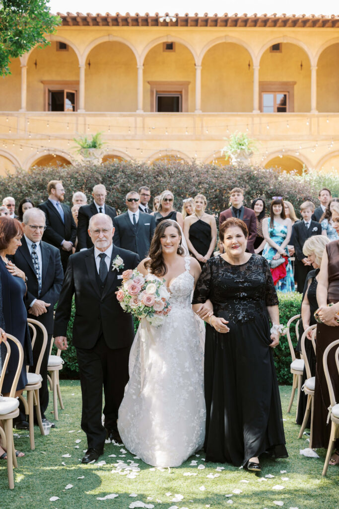 Villa Del Sol D'Oro Wedding Ceremony - A bride in a white gown holding a bouquet walks down an outdoor aisle, escorted by an older man and woman in formal attire. Guests seated and standing watch the procession; a yellow building with arches is in the background. Copyright - Lisa Leanne Photography, California