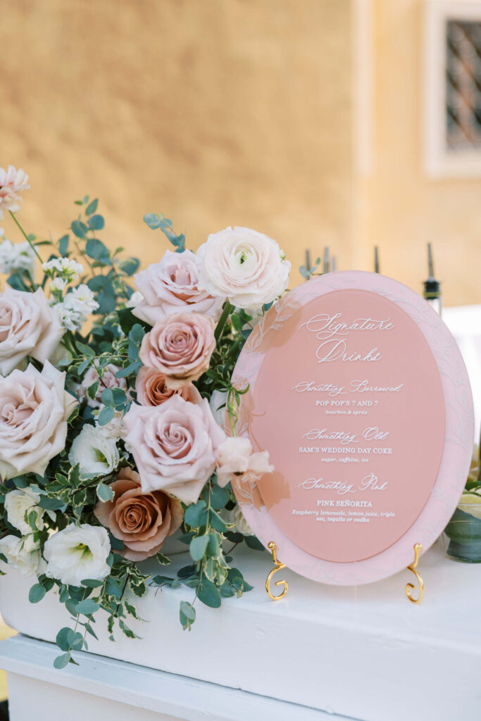 Cocktail Hour at Villa Del Sol D'Oro - A pink oval drinks menu with elegant white script stands on a gold holder, surrounded by an arrangement of blush and cream roses, eucalyptus, and greenery against a soft yellow background. Copyright - Lisa Leanne Photography, California
