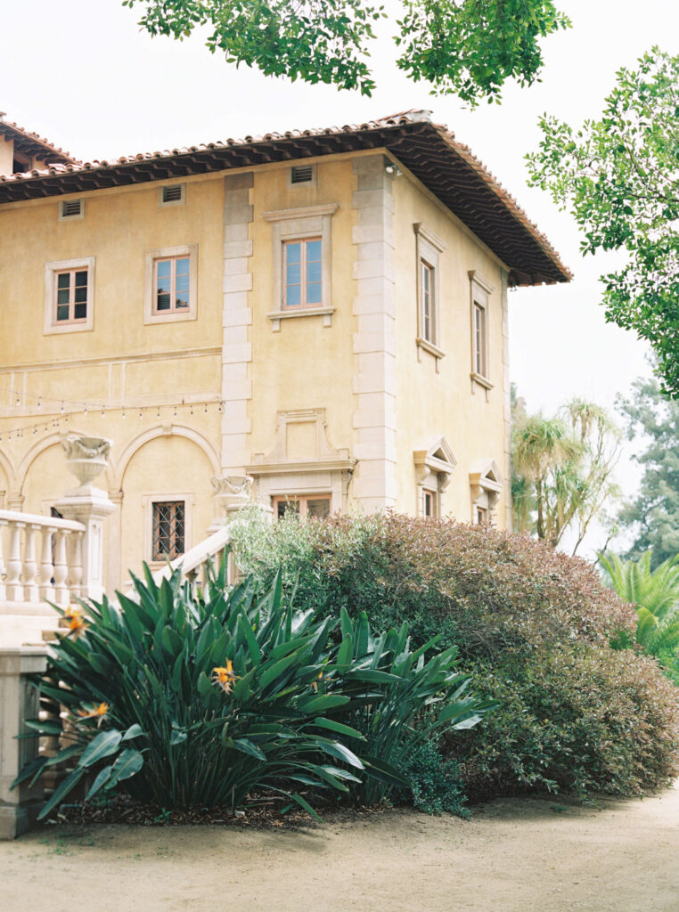 Villa Del Sol D'Oro Wedding - A Mediterranean-style villa with yellow walls, red-tiled roof, arched windows, and a stone staircase, surrounded by lush green plants and trees on a sunny day. Copyright - Lisa Leanne Photography, California