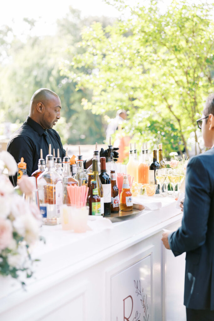 Cocktail Hour at Villa Del Sol D'Oro - A bartender stands behind an outdoor bar filled with bottles, mixers, and glasses, serving a guest in a suit on a sunny day. Green trees and soft light create a cheerful atmosphere. Copyright - Lisa Leanne Photography, California