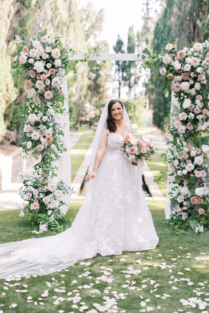 Villa Del Sol D'Oro Wedding Bridal Portrait - A bride in a white lace gown and veil stands smiling under a floral arch outdoors, holding a bouquet of pastel flowers; petals are scattered on the grass around her. Copyright - Lisa Leanne Photography, California