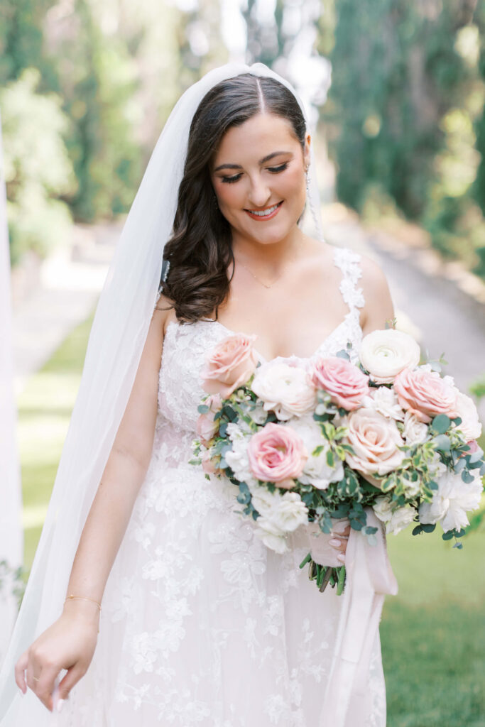 Villa Del Sol D'Oro Wedding Bridal Portrait - A bride in a white lace wedding dress holds a bouquet of pink and white roses, smiling softly, with greenery and a blurred outdoor background of trees and sunlight. Copyright - Lisa Leanne Photography, California