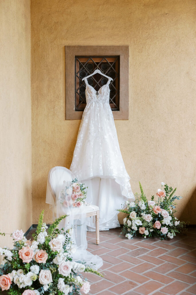 Villa Del Sol D'Oro Wedding - A white lace wedding dress hangs on a decorative window above a wooden chair draped with a white veil, surrounded by floral arrangements of white and blush pink roses and greenery against a tan stucco wall. Copyright - Lisa Leanne Photography, California