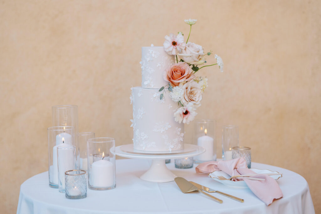 Villa Del Sol D'Oro Wedding Cake - A two-tier white wedding cake decorated with white lace-like icing and fresh flowers, placed on a white stand. Surrounded by lit candles, a cake server, and pink napkins on a round table. Copyright - Lisa Leanne Photography, California