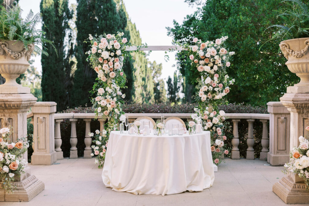Villa Del Sol D'Oro Reception Sweetheart Table - A small round table with a white tablecloth is set for two in front of a floral archway decorated with pink and white flowers on a stone terrace, surrounded by greenery and large planter urns. Copyright - Lisa Leanne Photography, California