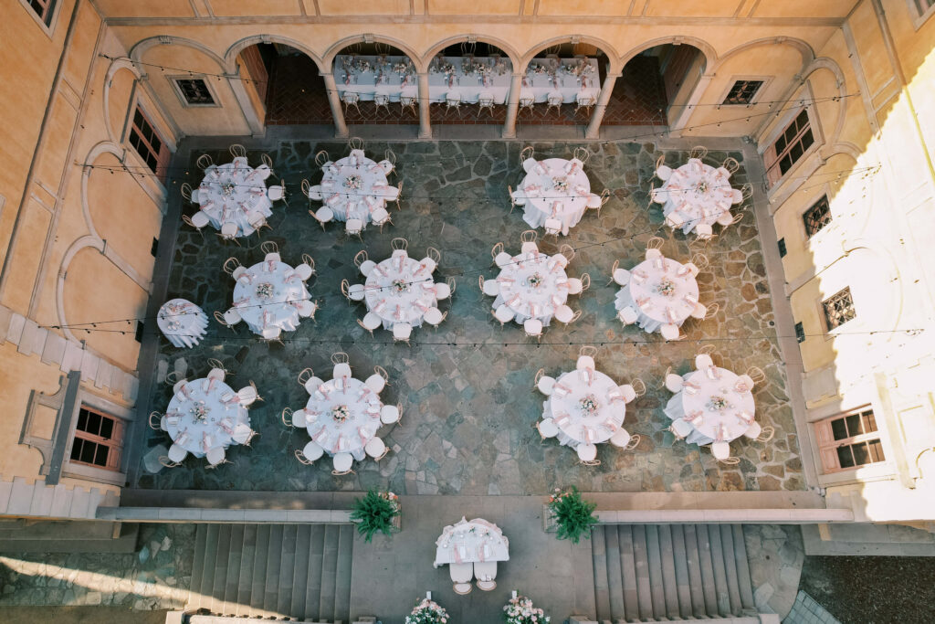 Villa Del Sol D'Oro Wedding Reception - Aerial view of an elegant outdoor courtyard set up for an event, with round tables covered in white tablecloths and chairs evenly spaced on a stone floor, surrounded by an arched building. Copyright - Lisa Leanne Photography, California