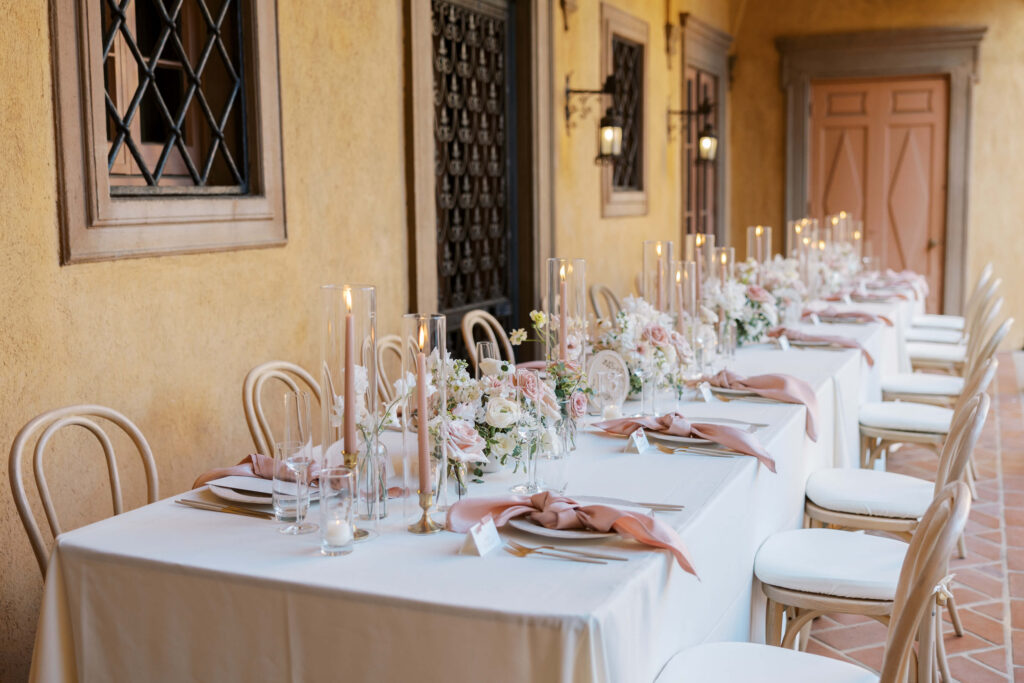 Villa Del Sol D'Oro Wedding Reception Setup - A long, elegant table set for a formal event with white linens, blush pink napkins, flowers, and tall candles. The table is surrounded by white chairs in a warm, outdoor Mediterranean-style corridor. Copyright - Lisa Leanne Photography, California