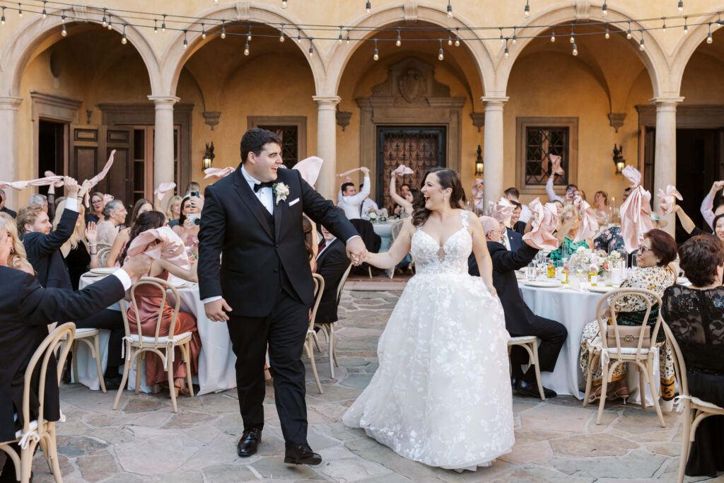 Villa Del Sol D'Oro Reception Grand Entrance - A bride and groom, holding hands and smiling, walk into an outdoor courtyard reception as guests seated at tables cheer and wave napkins in celebration. String lights hang above, and the setting is elegant with arches. Copyright - Lisa Leanne Photography, California
