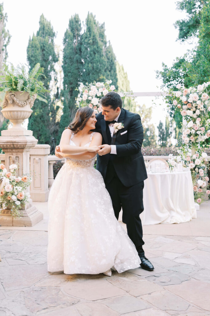 Villa Del Sol D'Oro Reception First Dance - A bride and groom share a joyful dance outdoors, surrounded by lush greenery and elegant floral arrangements, with the bride in a white gown and the groom in a black suit. Copyright - Lisa Leanne Photography, California