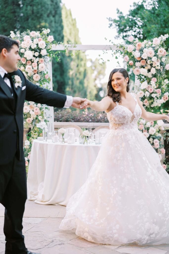 Villa Del Sol D'Oro Reception First Dance - A bride and groom dance outdoors in front of a table and lush floral arrangements. The bride wears a white lace gown and smiles as she twirls, holding the groom’s hand. The groom is in a black tuxedo. Copyright - Lisa Leanne Photography, California