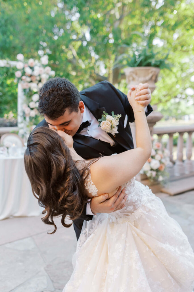 Villa Del Sol D'Oro Reception First Dance - A groom in a black tuxedo dips and kisses his bride in a white wedding dress outdoors, surrounded by flowers and greenery, during their wedding celebration. Copyright - Lisa Leanne Photography, California