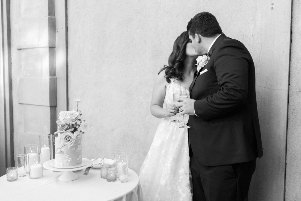 Villa Del Sol D'Oro Reception Cake Cutting - A bride and groom kiss while holding drinks beside a small, decorated wedding cake on a table with candles and flowers. The image is in black and white. Copyright - Lisa Leanne Photography, California
