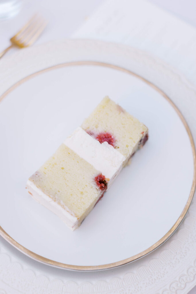 Villa Del Sol D'Oro Wedding - A slice of vanilla cake with white frosting and raspberries inside, served on a white plate with a gold rim. Copyright - Lisa Leanne Photography, California