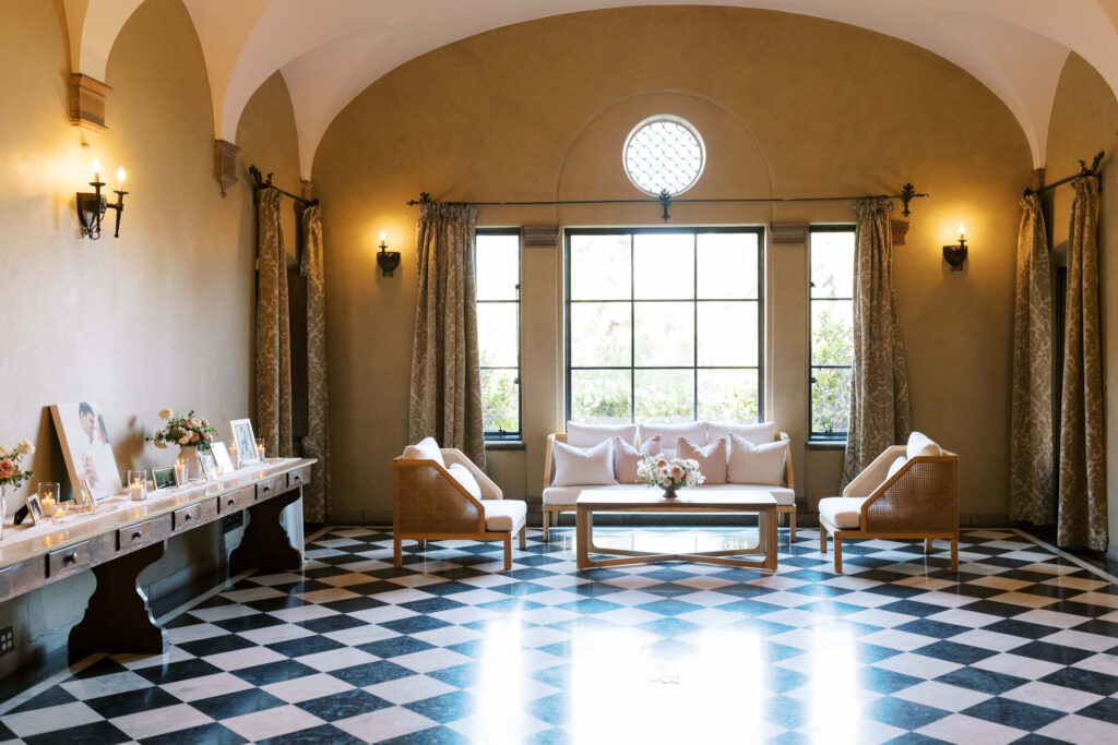 Villa Del Sol D'Oro - Elegant sitting room with black-and-white checkered floor, neutral walls, arched ceiling, large windows with curtains, and cozy seating around a coffee table topped with flowers. Decor items line a long side table. Copyright - Lisa Leanne Photography, California