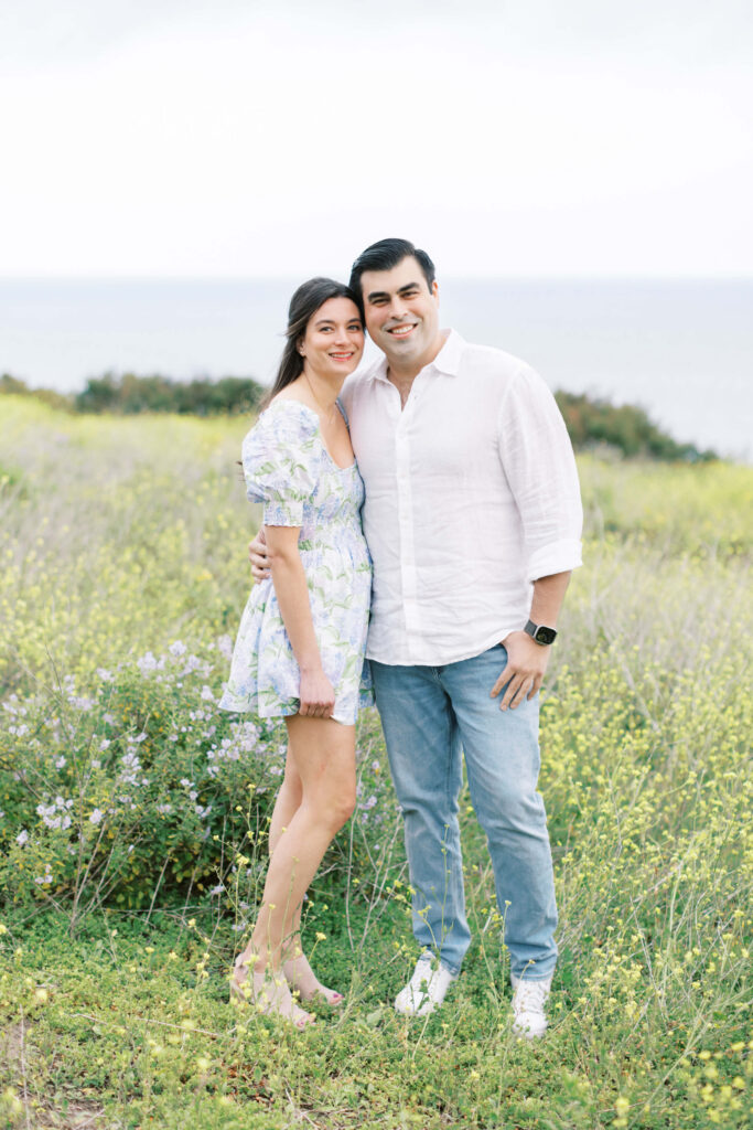 Malibu Beach Engagement Session - A smiling couple stands close together in a grassy field with wildflowers, with the ocean and a cloudy sky in the background. The woman wears a floral dress; the man wears a white shirt and jeans. Copyright - Lisa Leanne Photography, California