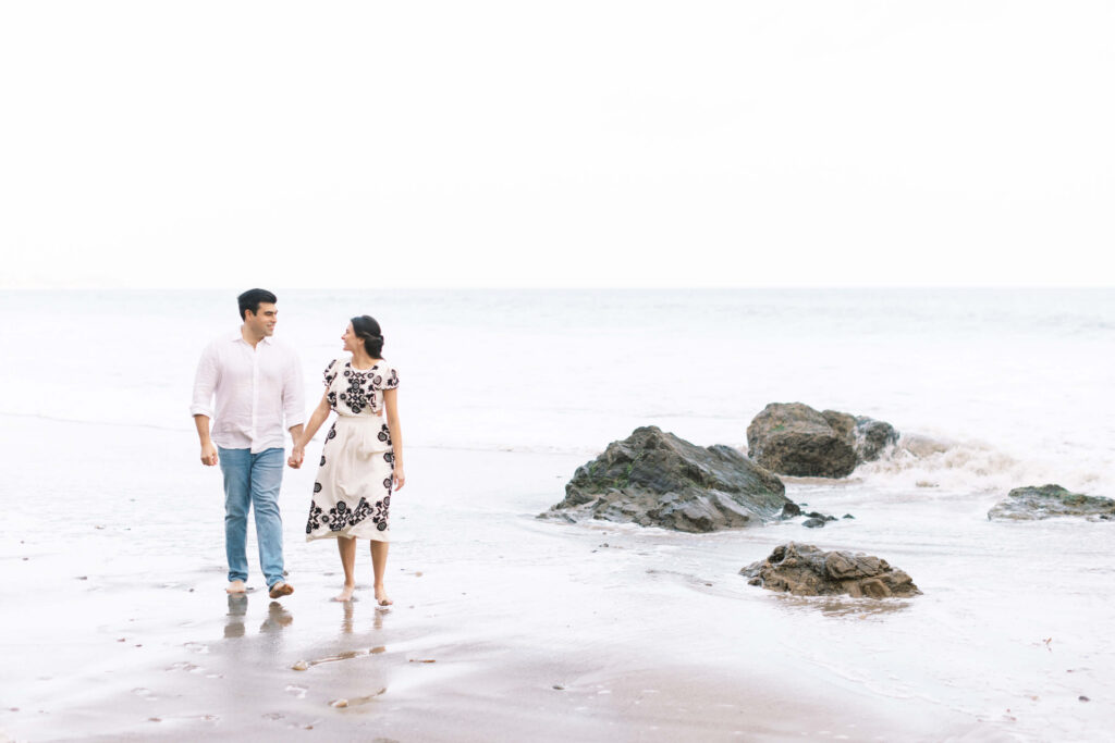 El Matador Beach Engagement Session - A couple walks hand-in-hand along a sandy beach near rocks, looking at each other and smiling. The ocean waves and overcast sky create a calm, serene atmosphere. Copyright - Lisa Leanne Photography, California