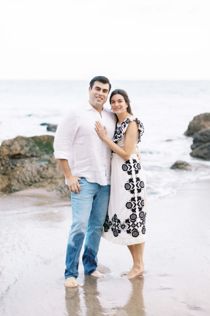 El Matador Beach Engagement Session - A couple stands barefoot on a sandy beach near rocks, smiling at the camera. The man wears a white shirt and jeans, while the woman wears a white dress with black floral patterns. The ocean waves are in the background. Copyright - Lisa Leanne Photography, California