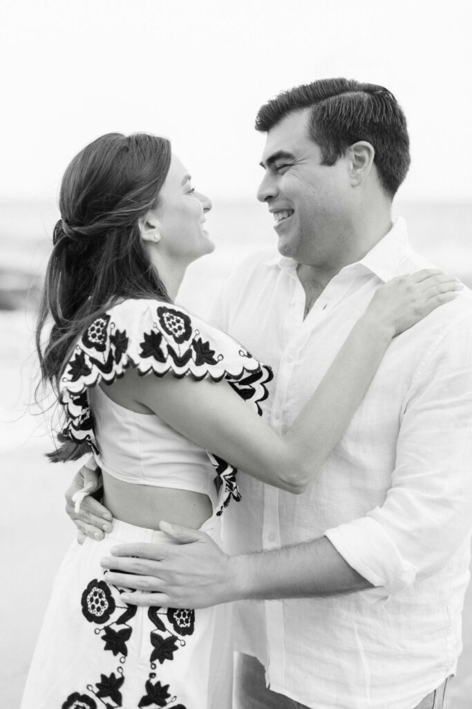 El Matador Beach Engagement Session - A couple smiles at each other while embracing on the beach. The woman wears a white dress with black embroidery, and the man wears a white shirt. The photo is in black and white. Copyright - Lisa Leanne Photography, California
