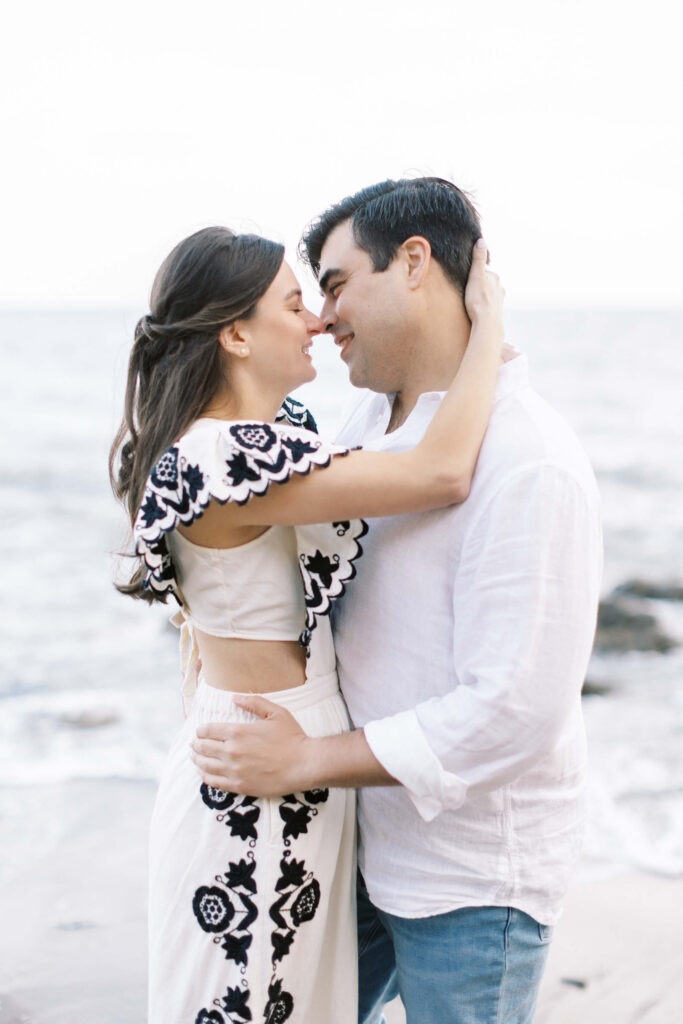 El Matador Beach Engagement Session - A couple stands close together on a beach, smiling and touching noses. The woman has long brown hair and wears a white and black patterned dress; the man wears a white shirt and jeans. The ocean is in the background. Copyright - Lisa Leanne Photography, California